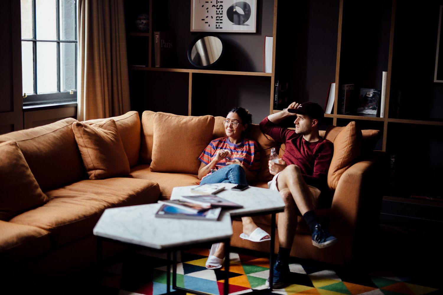 A couple relaxes together on a brown leather couch in a dimly lit room.