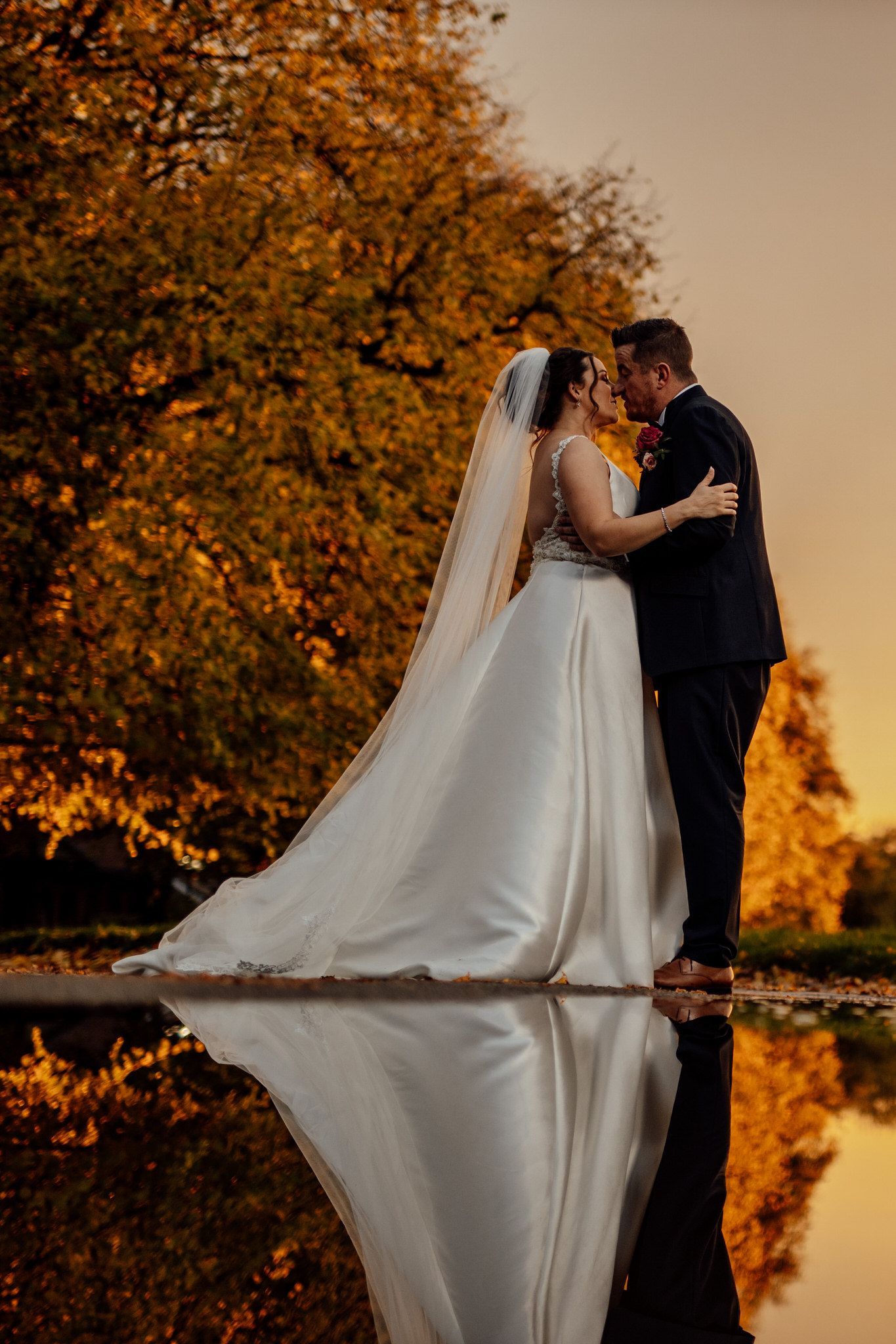 Dramatic sunset wedding portraits with flowing white dress against autumn foliage.