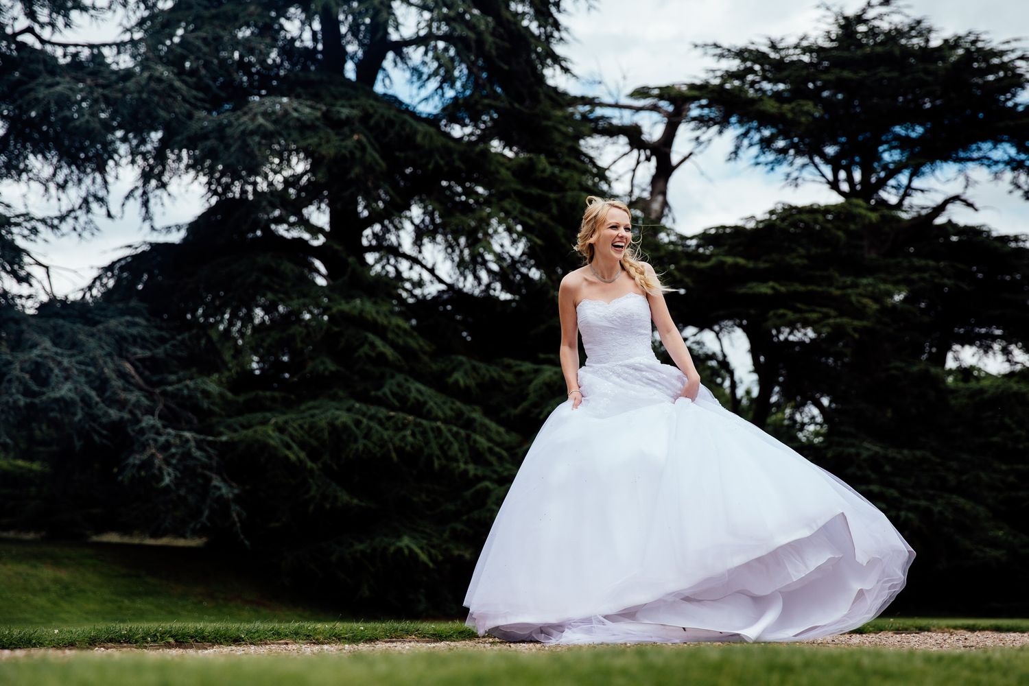 Bride in white gown twirls on manicured lawn with tall trees in background.