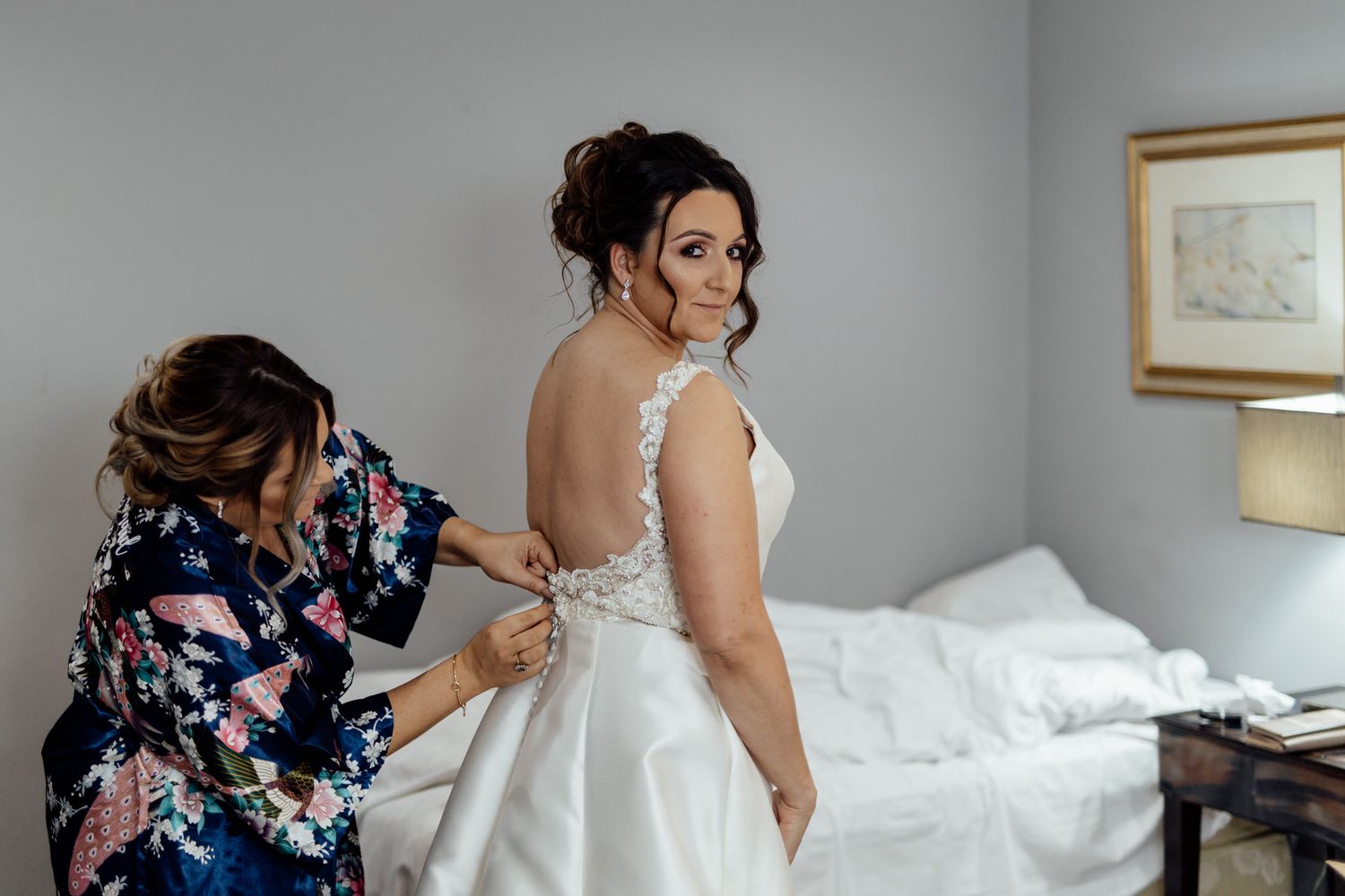 Final dress preparations in a bright hotel room with elegant white wedding gown.