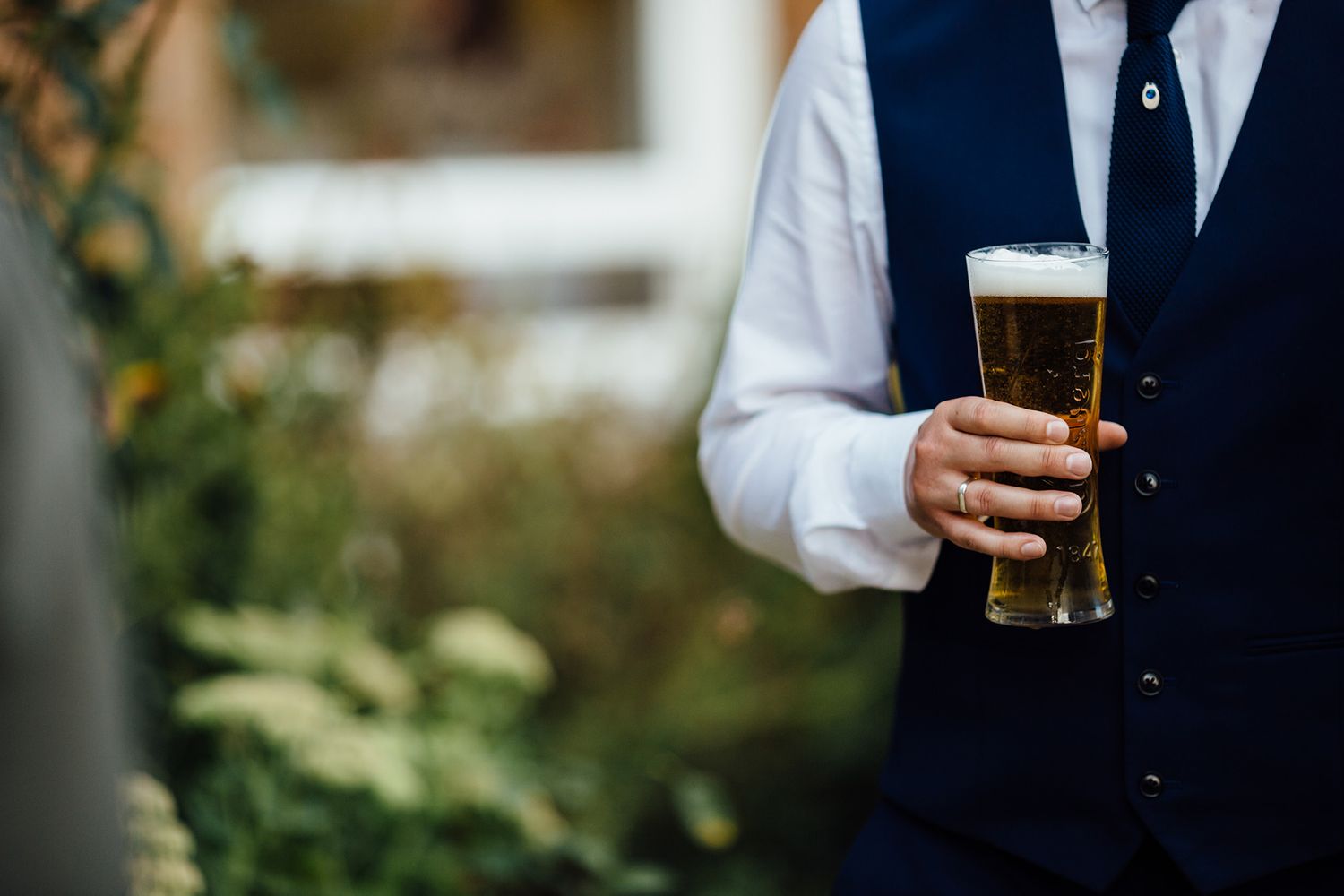 A hand holding a glass of dark beer wearing formal attire at an outdoor event.