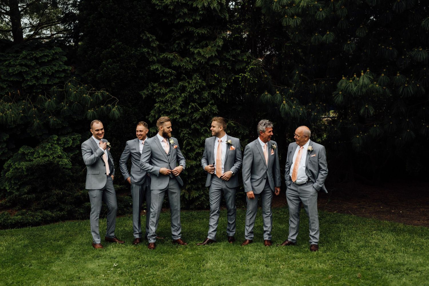 Groomsmen in matching gray suits pose together on a grassy lawn during a wedding celebration.