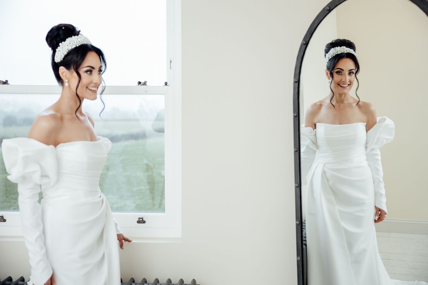 A bride in an elegant off-shoulder white dress poses in a bright room with natural lighting.