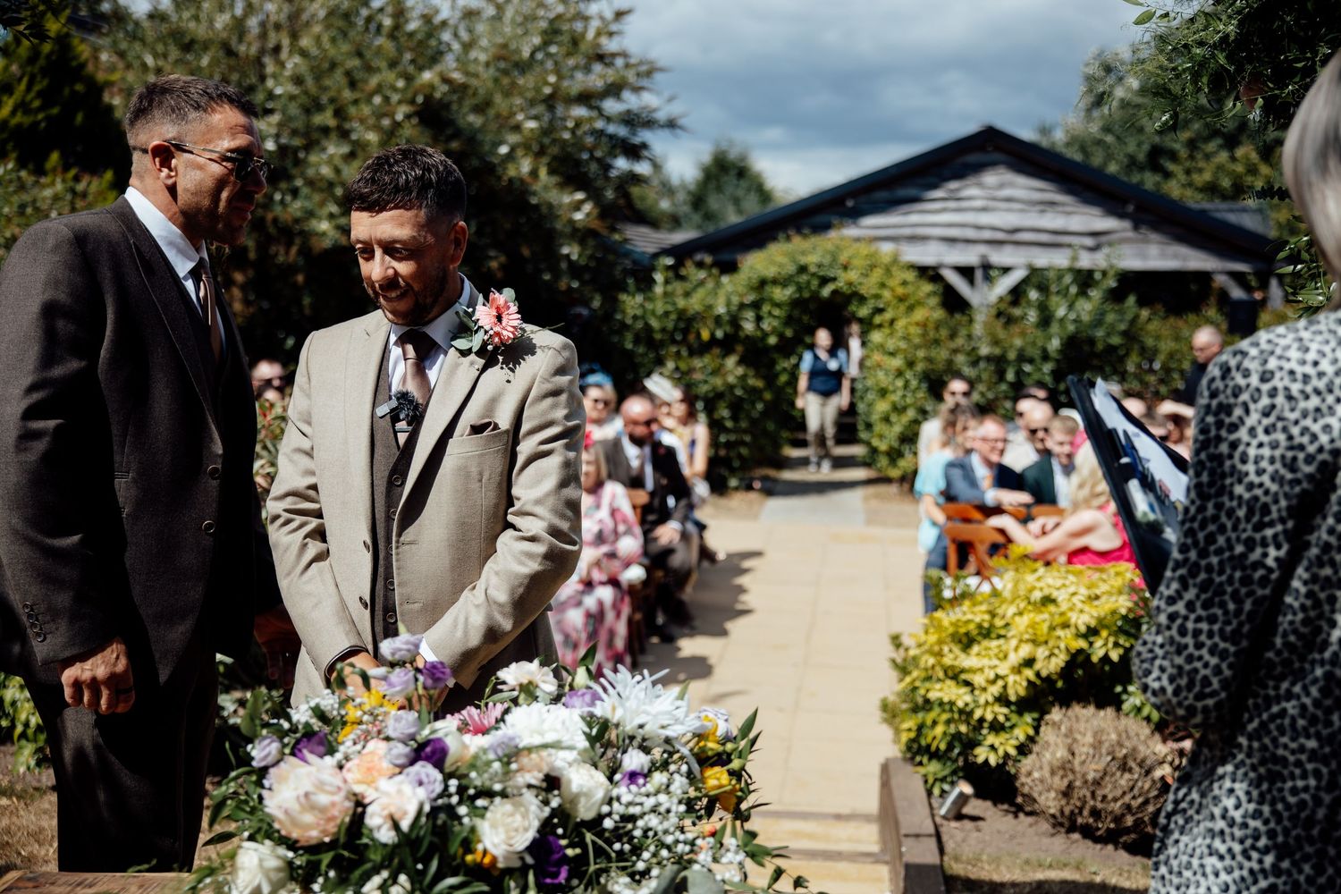 Groom in tan suit standing at outdoor wedding ceremony aisle with floral arrangements and seated guests.