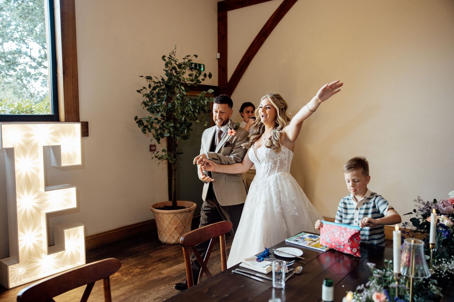 Bride in white dress gestures excitedly inside a rustic wedding venue with illuminated letter decor.