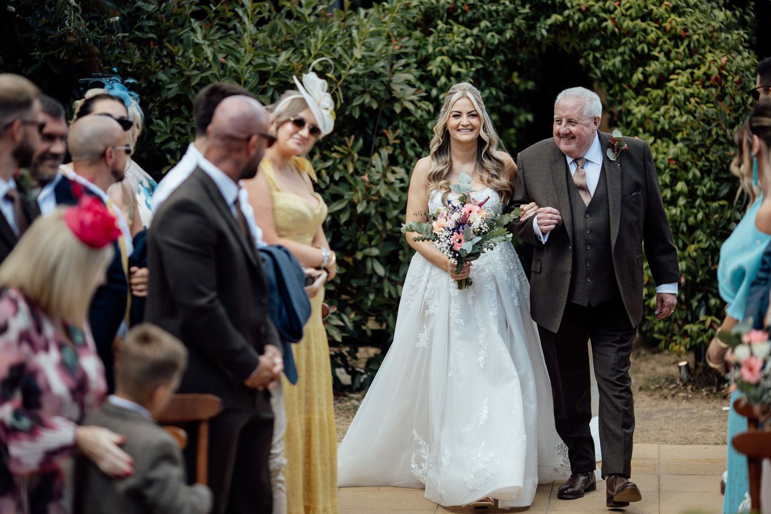 Radiant bride in lace wedding gown walks down outdoor aisle with father among smiling wedding guests.