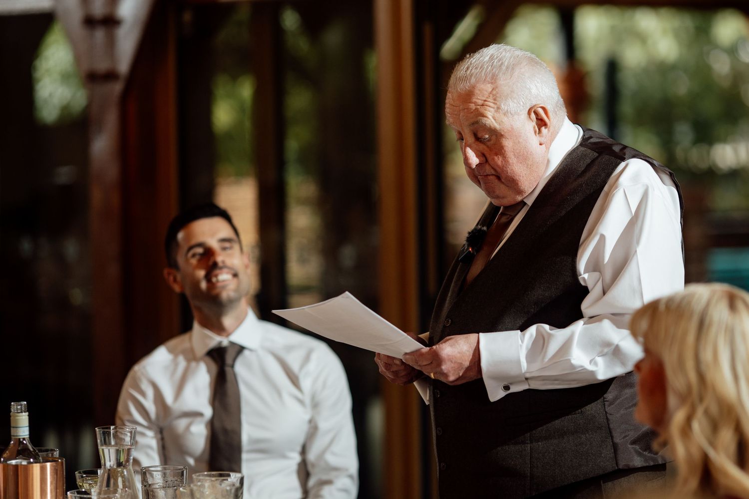 Elderly man in vest reads speech at formal dinner while smiling guest listens in elegant restaurant setting.