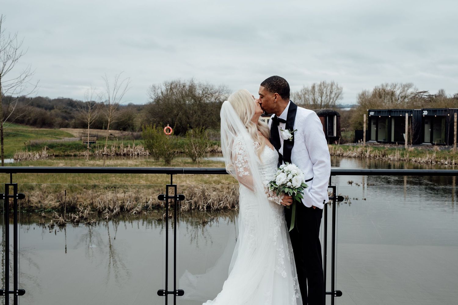 Interracial bride and groom sharing romantic kiss by pond at outdoor wedding venue.