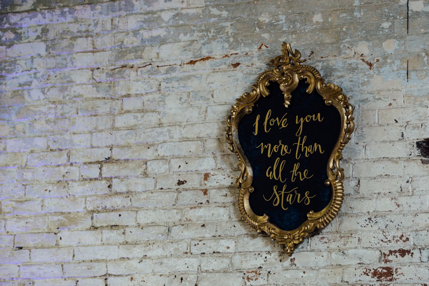 An ornate gold mirror frame with romantic text hangs on a weathered brick wall.
