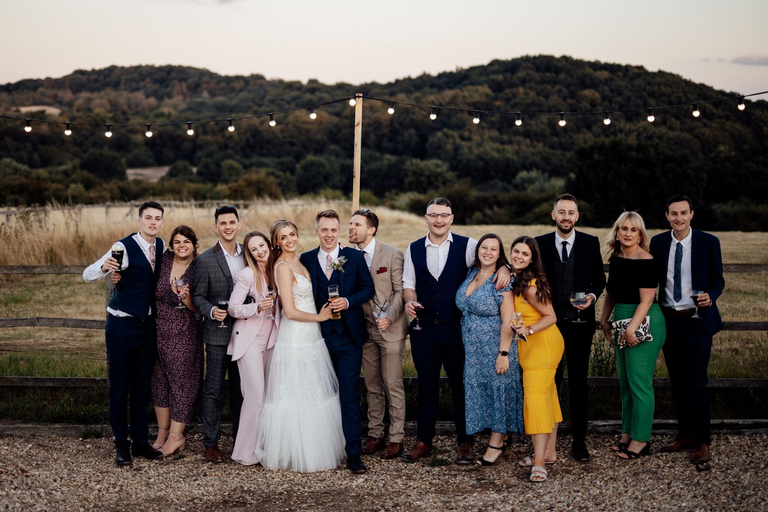Wedding party poses together outdoors at dusk with string lights and hills in background.