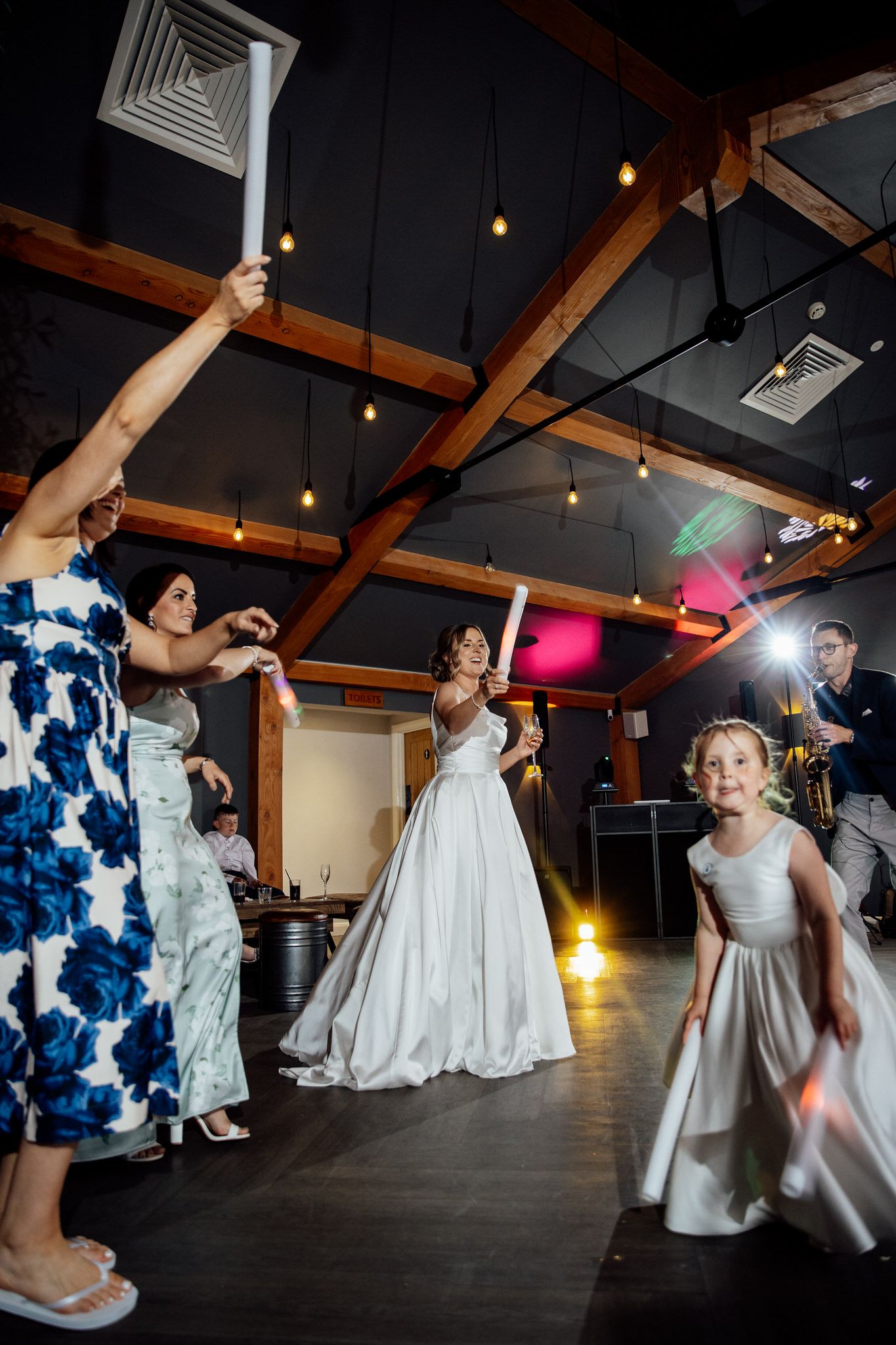 Wedding guests dancing and celebrating on a dark dance floor with exposed wooden beams and colorful lighting.