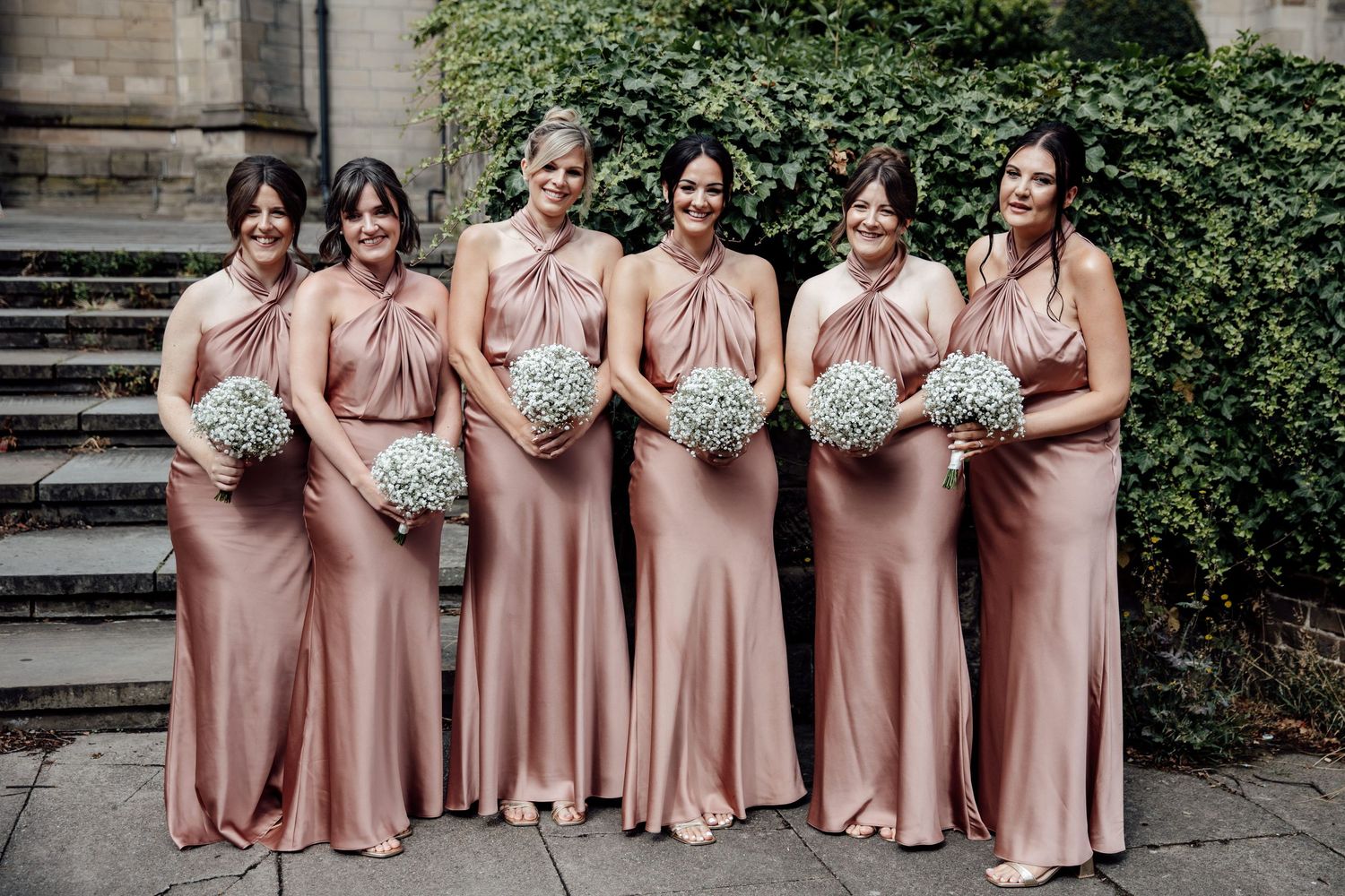 Bridesmaids wearing matching dusty rose satin dresses pose together on stone steps.