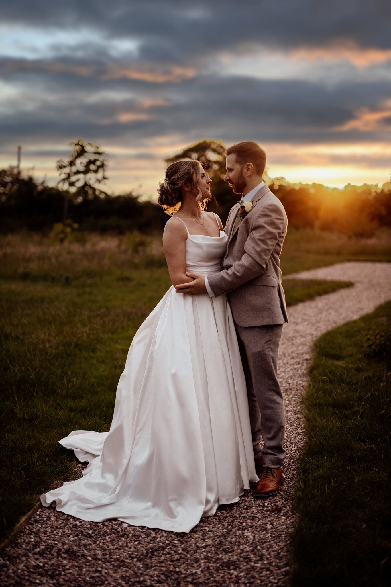 Couple embraces on a gravel path during golden hour with dramatic cloudy sunset sky.