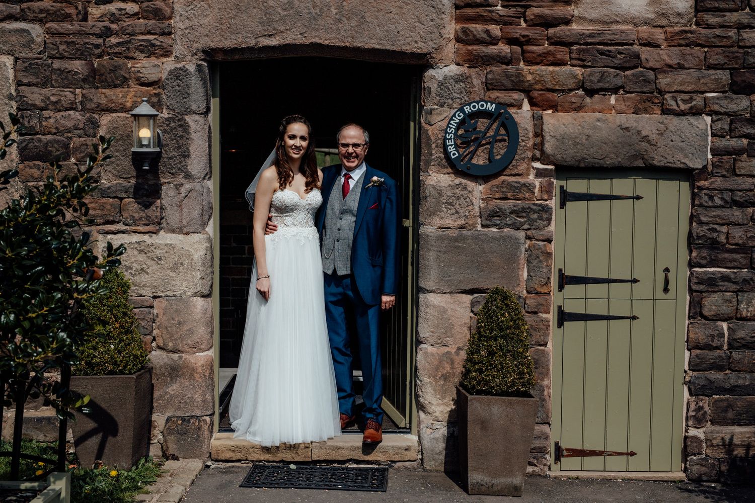 Newlyweds pose in doorway of rustic stone building with green shutters.