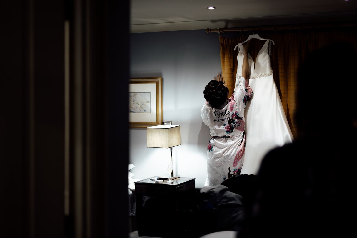 Silhouette of wedding preparation with dress hanging in hotel room window light.