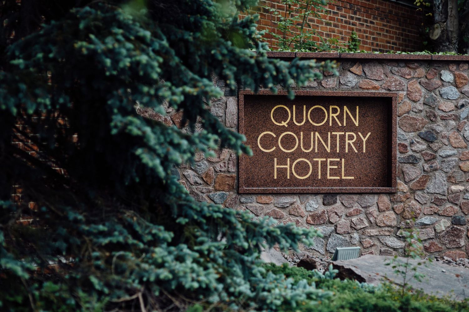 Rustic stone wall with wooden sign for Quorn Country Hotel nestled among greenery.