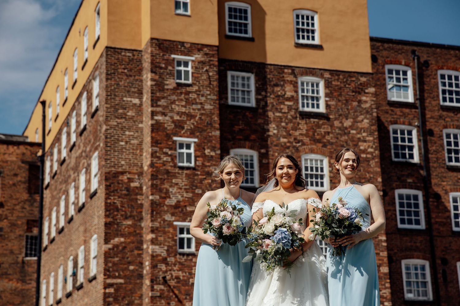 Bridal party in light blue dresses poses in front of historic brick building with bouquets.