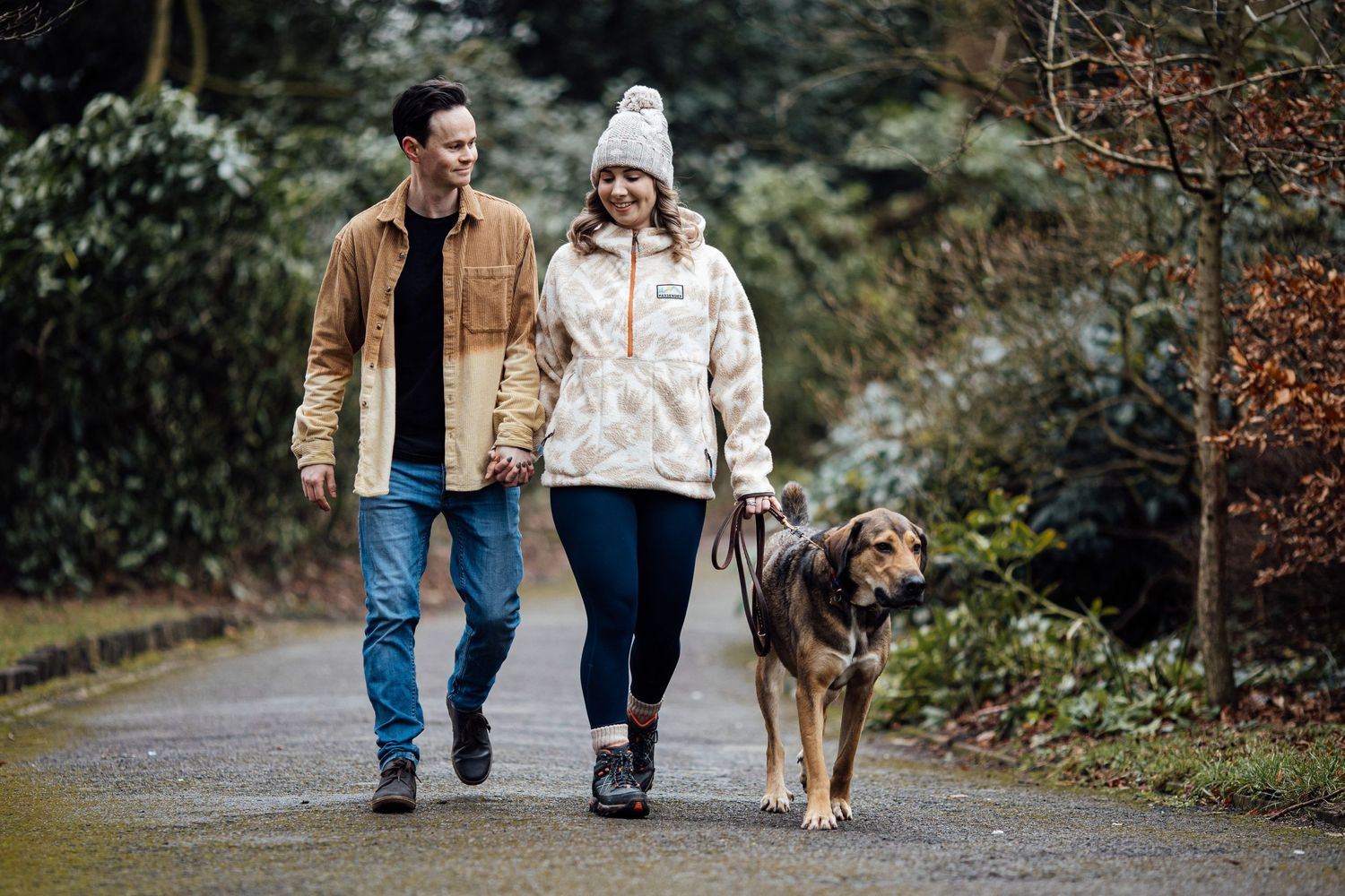Couple walking their dog along a wooded path during a casual outdoor photo session.