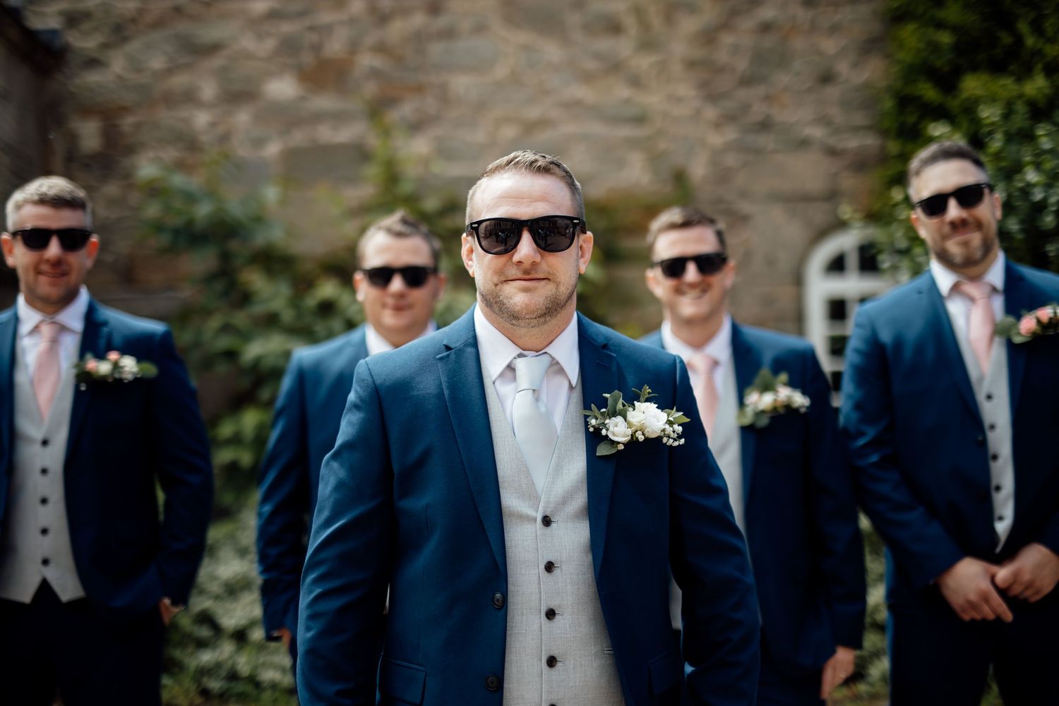Groomsmen in navy suits and sunglasses pose for formal wedding photos.