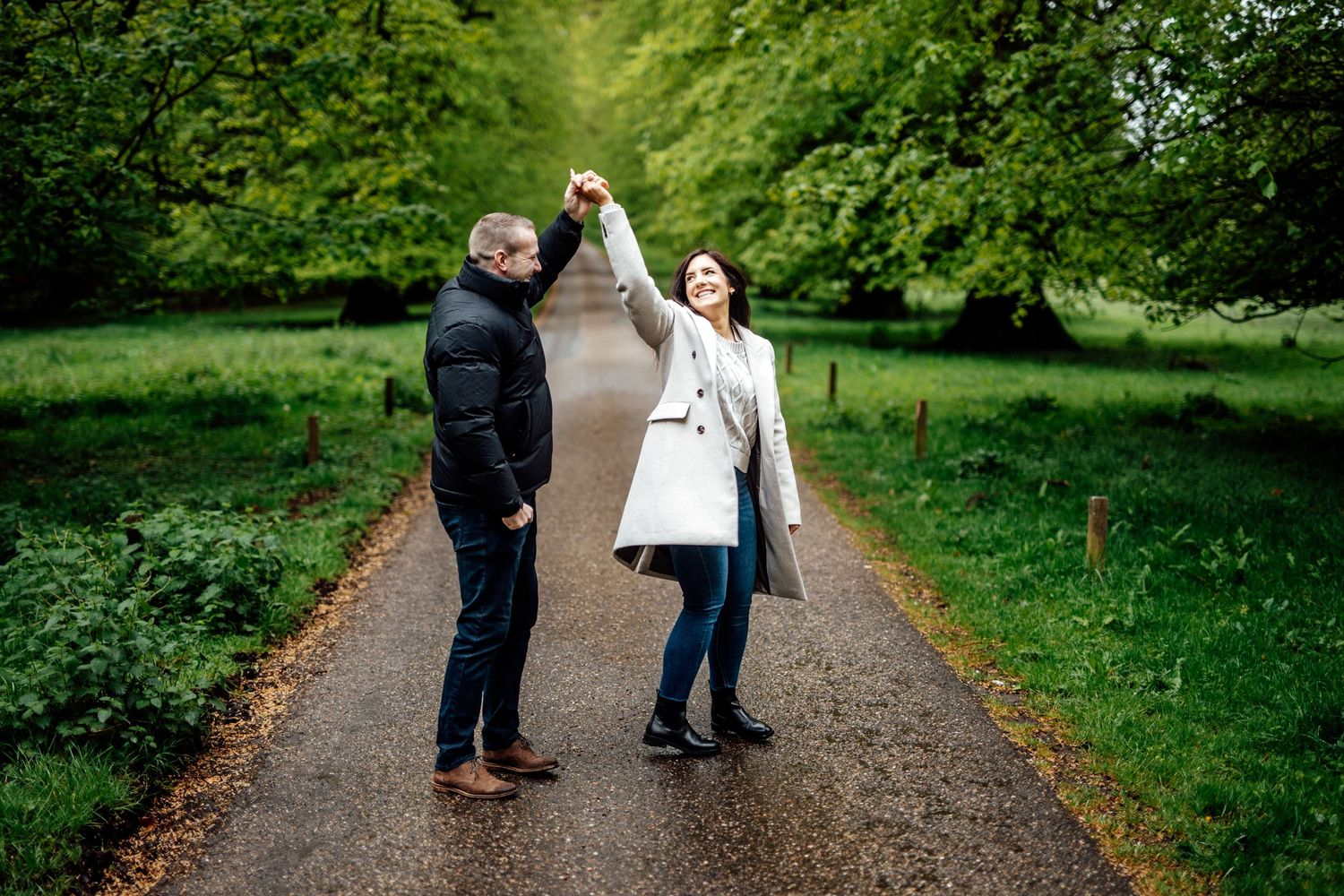 A couple dances playfully on a park path surrounded by green foliage.