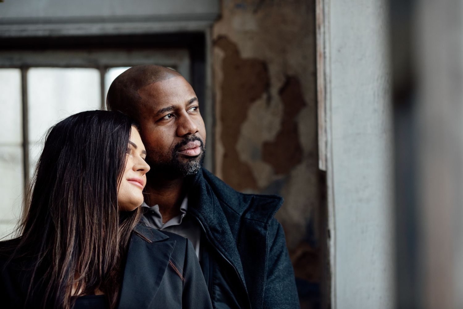 A couple in formal attire share an intimate moment while standing against a rustic wall with natural lighting.