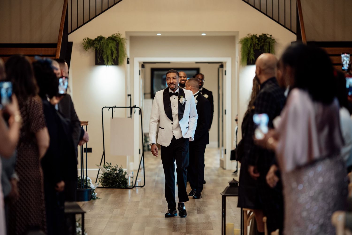 Two grooms walk down the wedding aisle together as guests capture the moment on their phones.