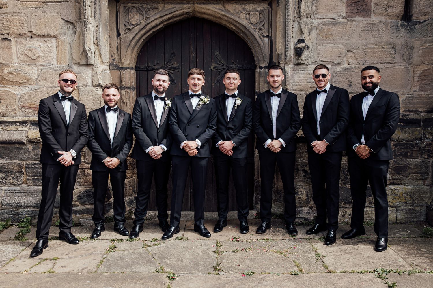 Groomsmen in black tuxedos pose against rustic stone wall backdrop.
