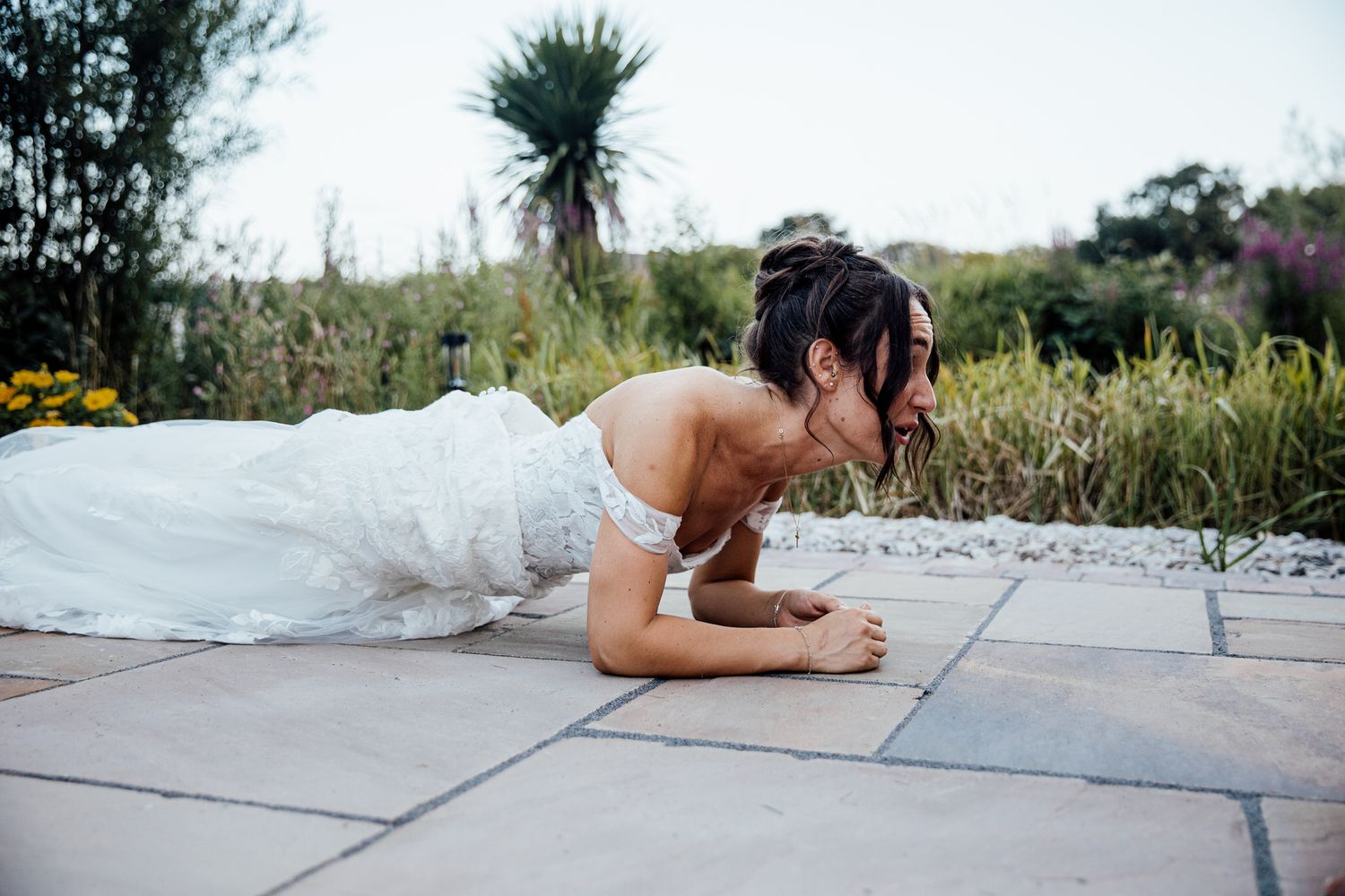 A bride in a white wedding dress does a playful plank pose on stone pavers in a garden.