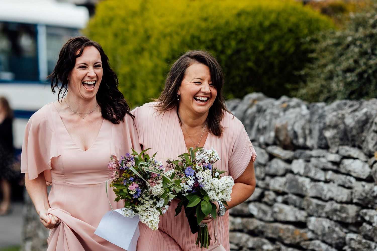 Two bridesmaids in blush pink dresses share a joyful laugh while holding bouquets.