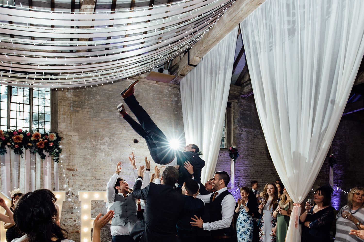 Wedding guests celebrate as someone is lifted into the air during a reception in an industrial venue with string lights.