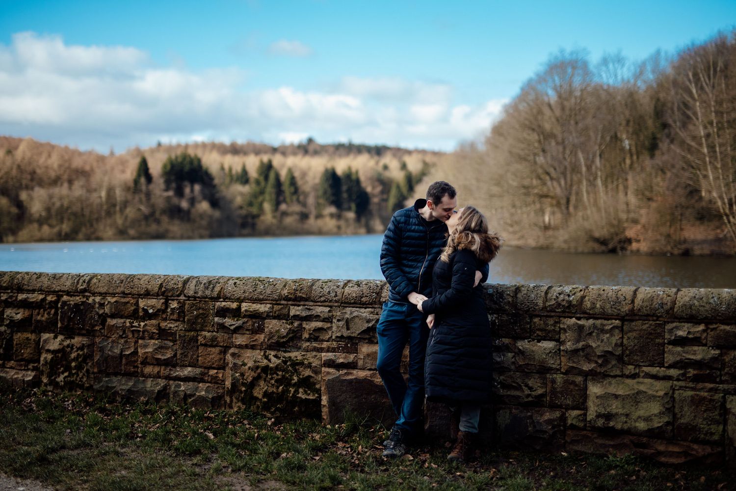 Couple sits together on a stone wall overlooking a scenic reservoir on a bright winter day.