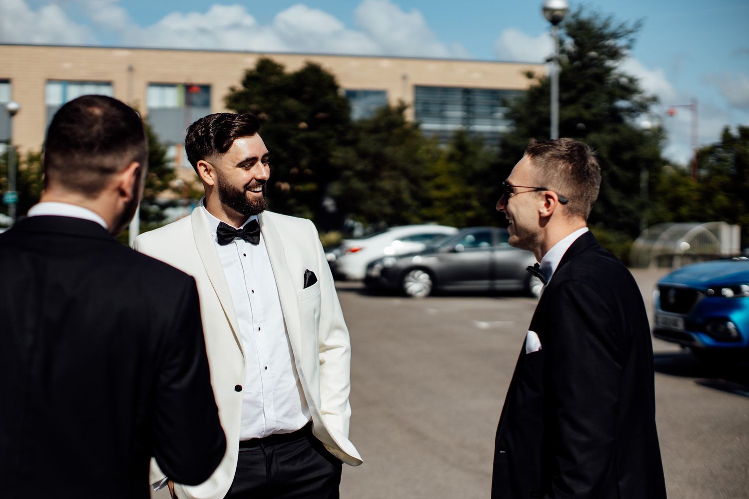 Groomsmen in white tuxedos and black bowties gathered outside on a sunny day.
