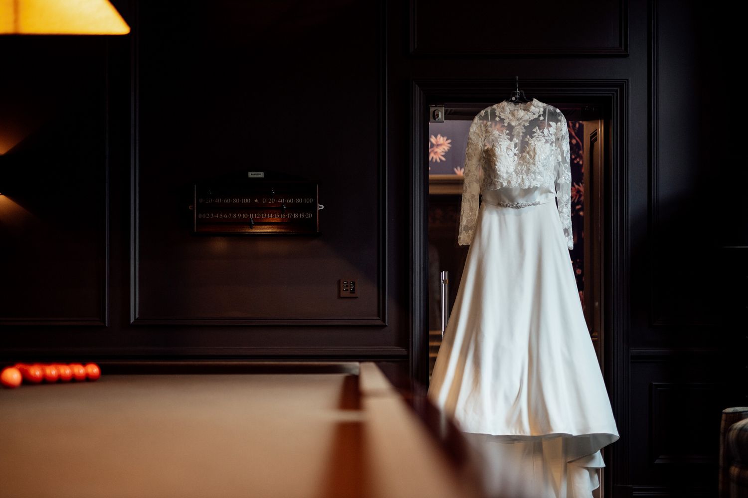 An elegant white wedding dress with lace details hanging in a dimly lit room.