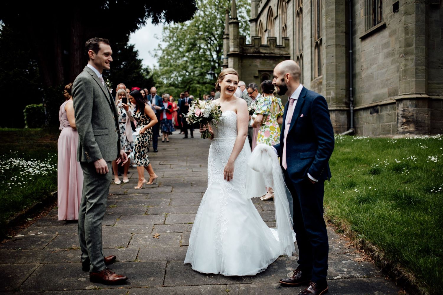 Wedding party walking along stone church pathway with guests looking on during outdoor ceremony.