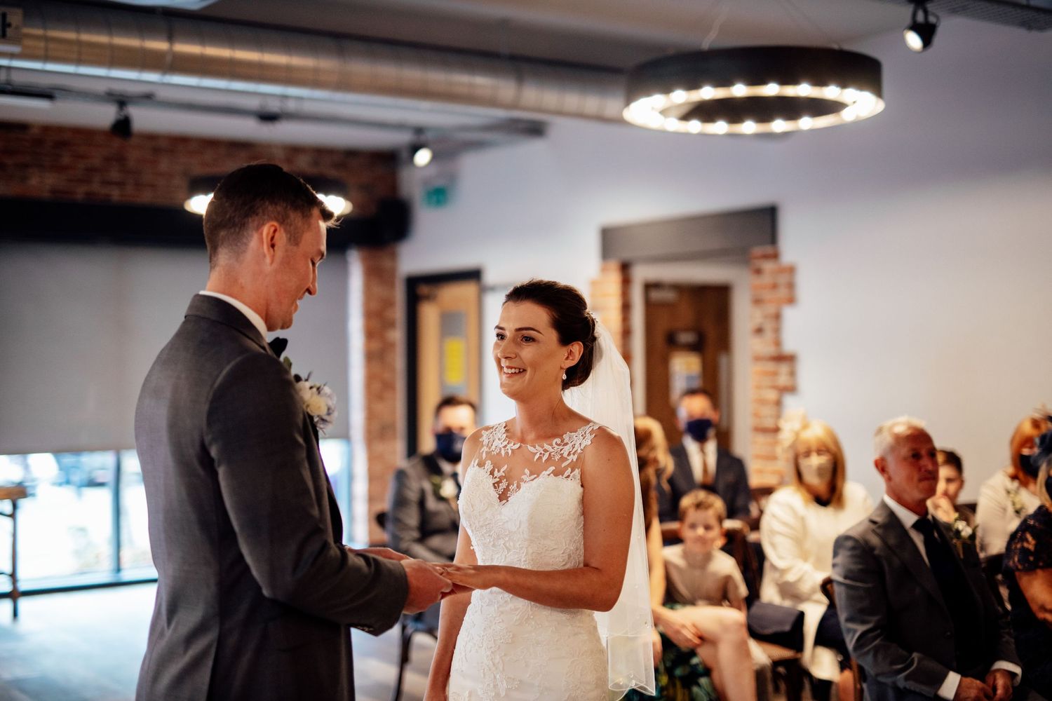 A couple exchanges wedding vows during an indoor ceremony with guests seated in the background.