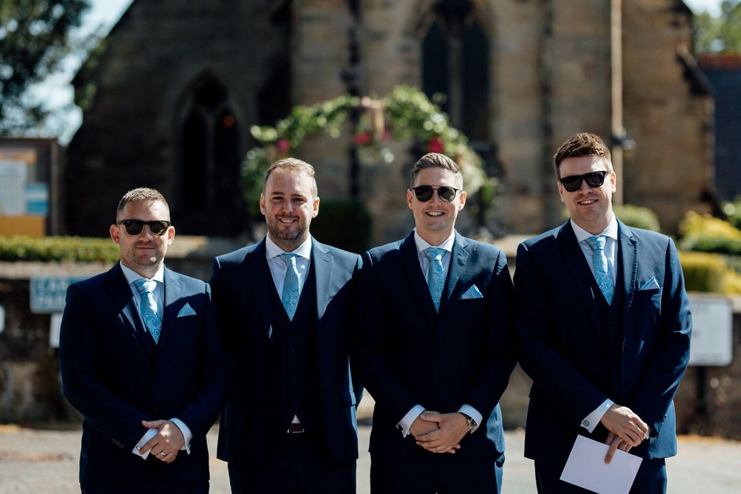 Group of groomsmen in dark suits and sunglasses posing outdoors.