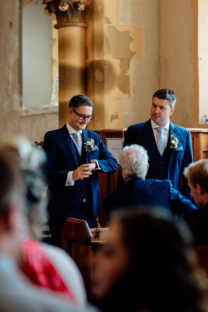 Men in navy suits share a laugh during church ceremony while seated in wooden pews.