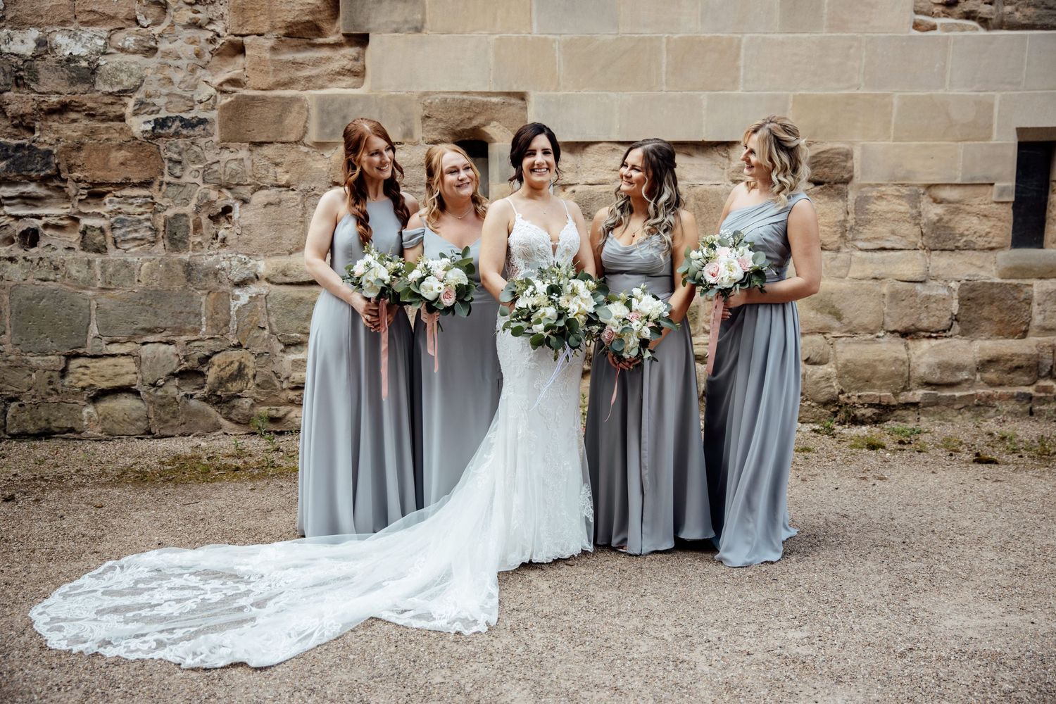 Bridal party in elegant gray dresses poses with bouquets against stone wall.