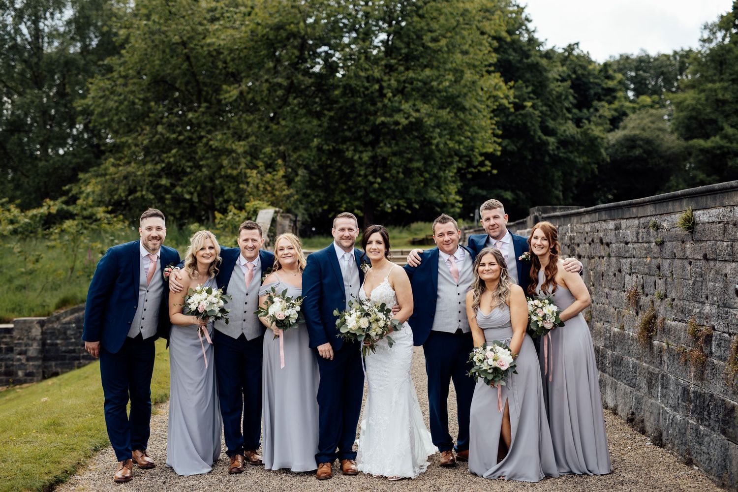 Wedding party group photo with bridesmaids and groomsmen in gray attire against stone wall backdrop.