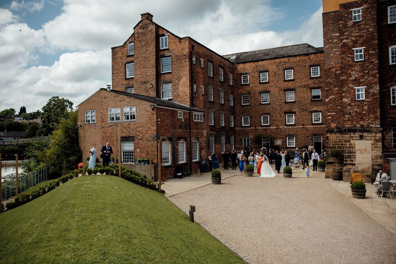 Historic brick mill building with wedding guests gathered outside on a sunny day.