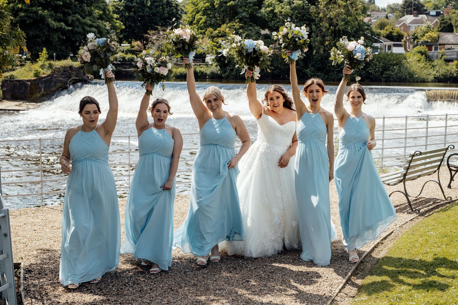 Bridesmaids in light blue dresses posing by waterfront with bride in white.