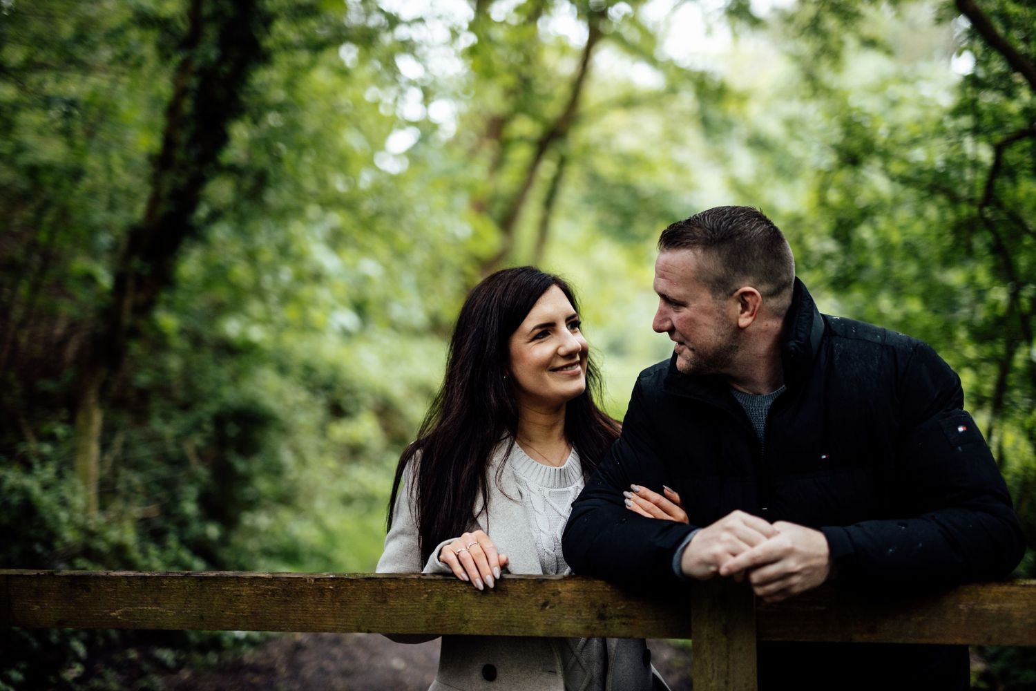 A couple sits close together on a wooden bench surrounded by trees and natural foliage.