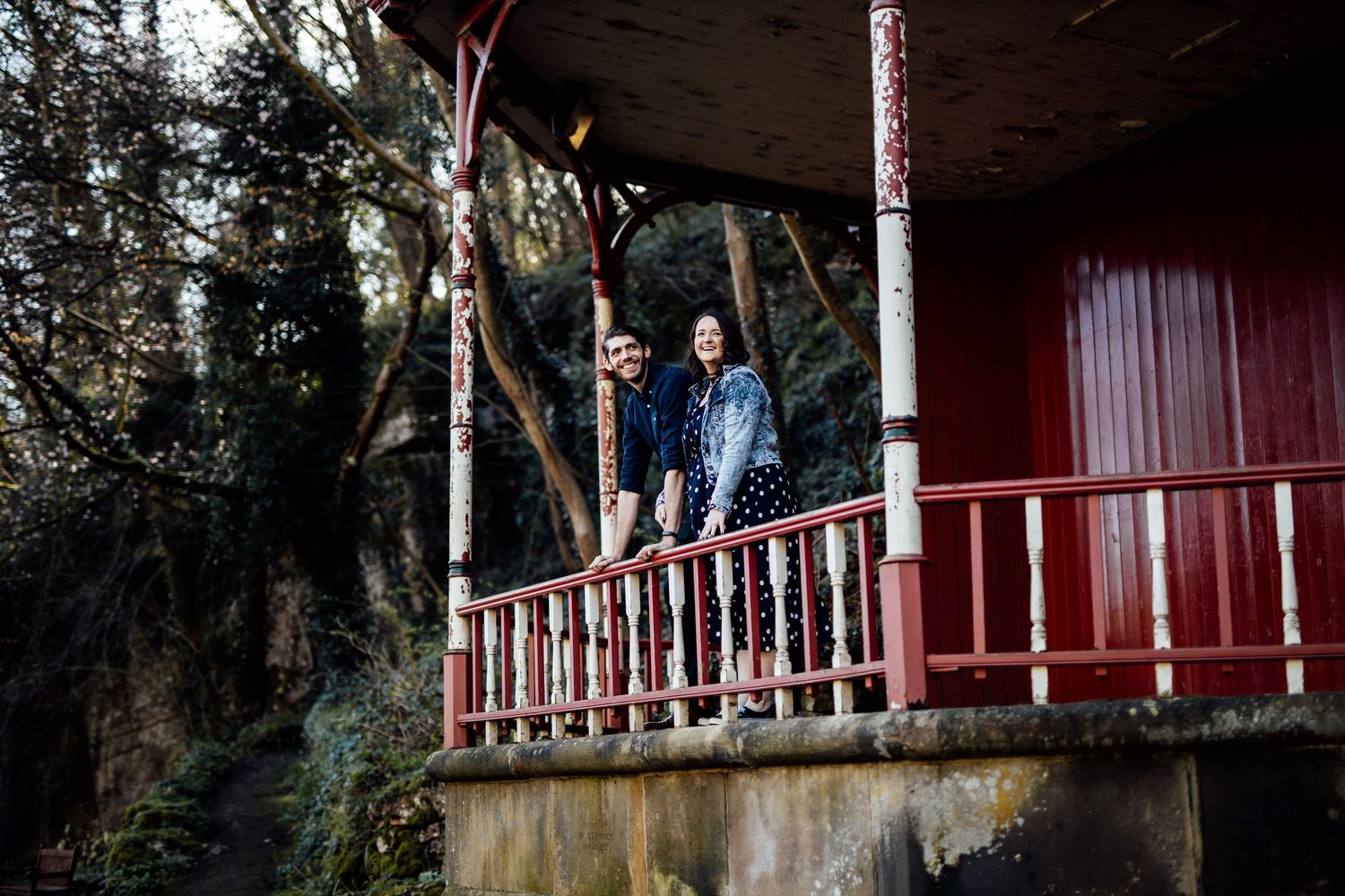 A covered walkway with wooden beams and red painted details extends through a wooded area.