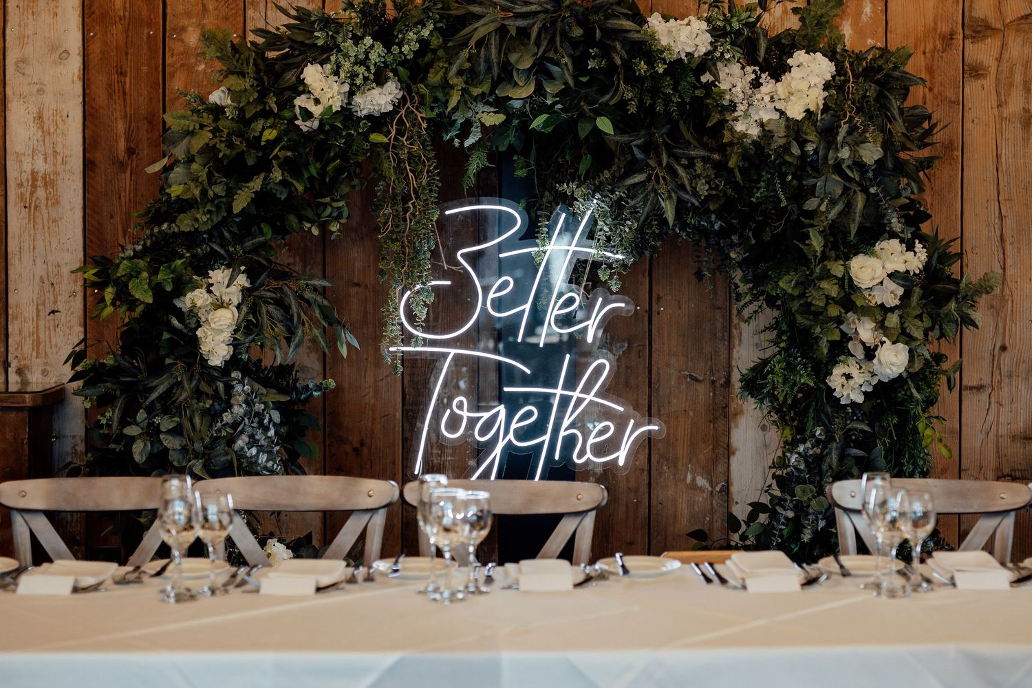 Rustic wooden wall decorated with floral arrangements and illuminated neon text signs.