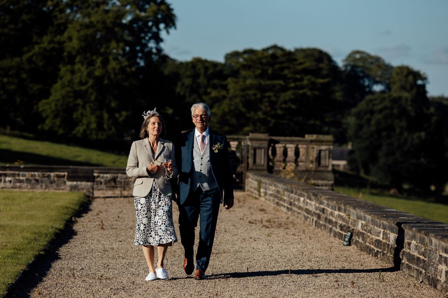 A couple walks along a scenic stone wall pathway surrounded by lush greenery on a sunny day.