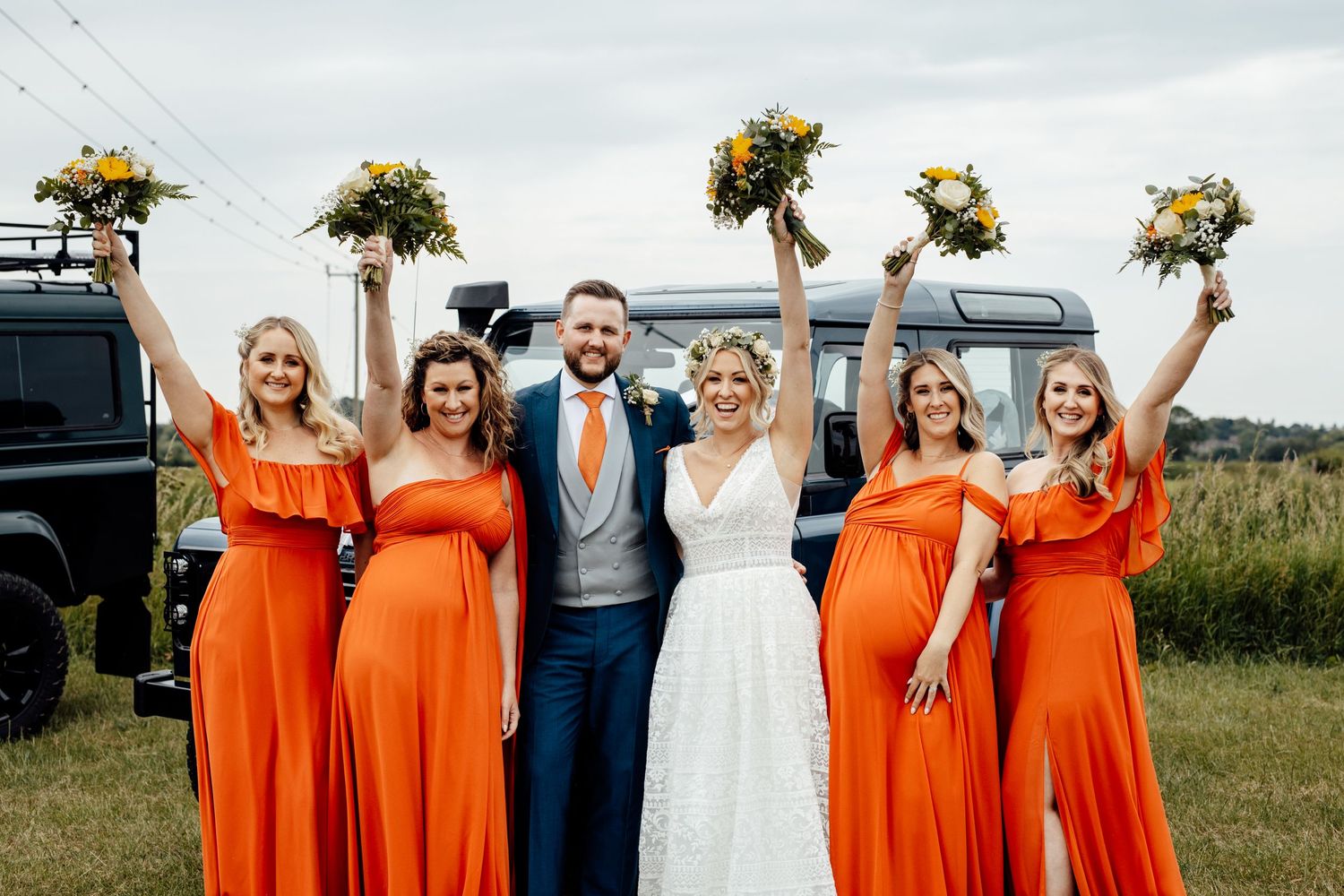 Wedding party poses with bright orange bridesmaid dresses and sunflowers against vintage Land Rovers.
