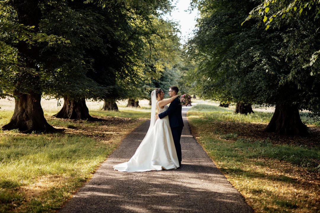 Wedding party gathers under large trees in outdoor park setting.