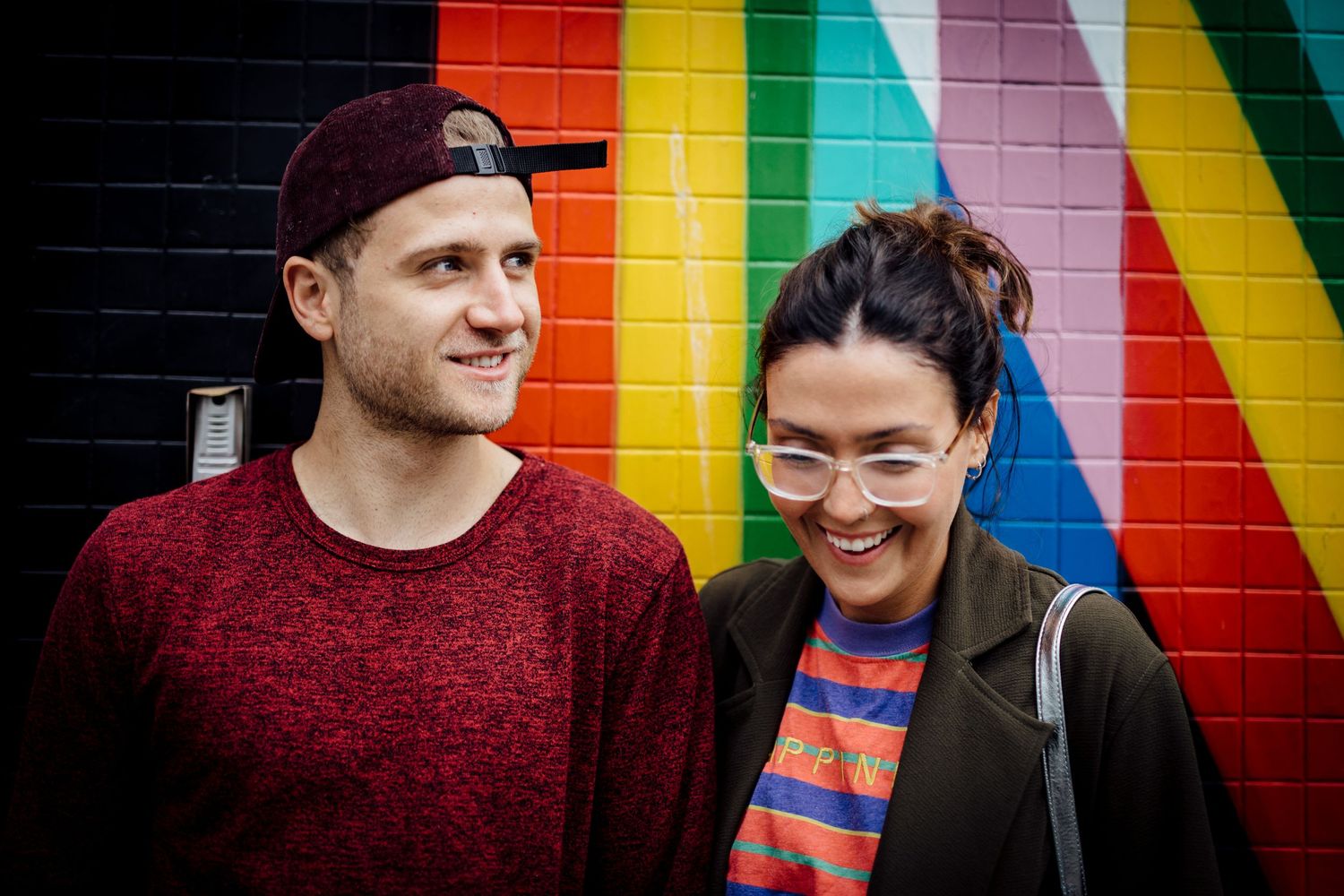 Two people laughing together in front of colorful striped wall mural.