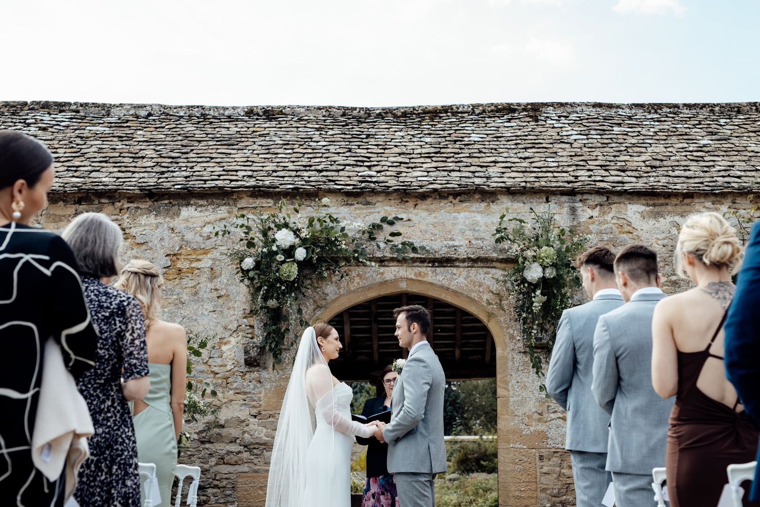 A wedding ceremony takes place in front of a rustic stone archway with guests gathered around.