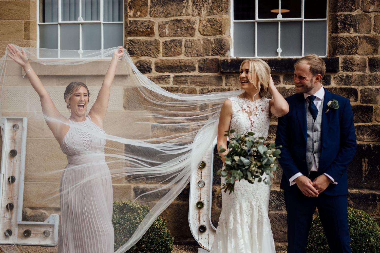 Wedding celebration group photo with flowing veil against stone building backdrop.